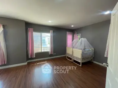 Spacious bedroom with wooden flooring and large window, featuring a cozy crib with pink curtains.