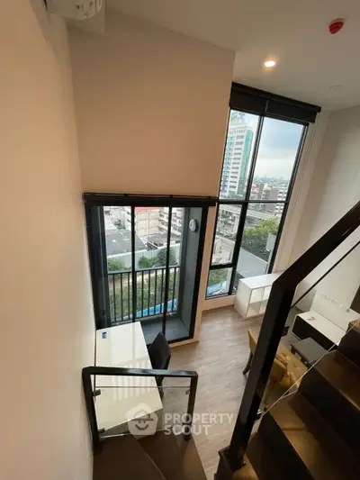 Modern duplex apartment with large windows and city view, featuring a stylish staircase and workspace.