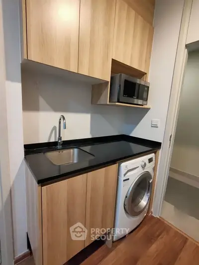 Modern compact kitchen with wooden cabinets, washing machine, and microwave.