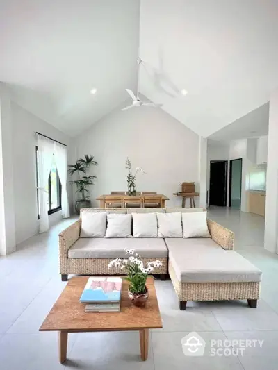 Spacious and airy living room with high vaulted ceiling, large wicker sectional sofa, and minimalist wooden coffee table, opening up to a bright dining area.