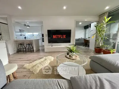Modern living room with open kitchen and stylish decor, featuring a large TV and cozy seating area.