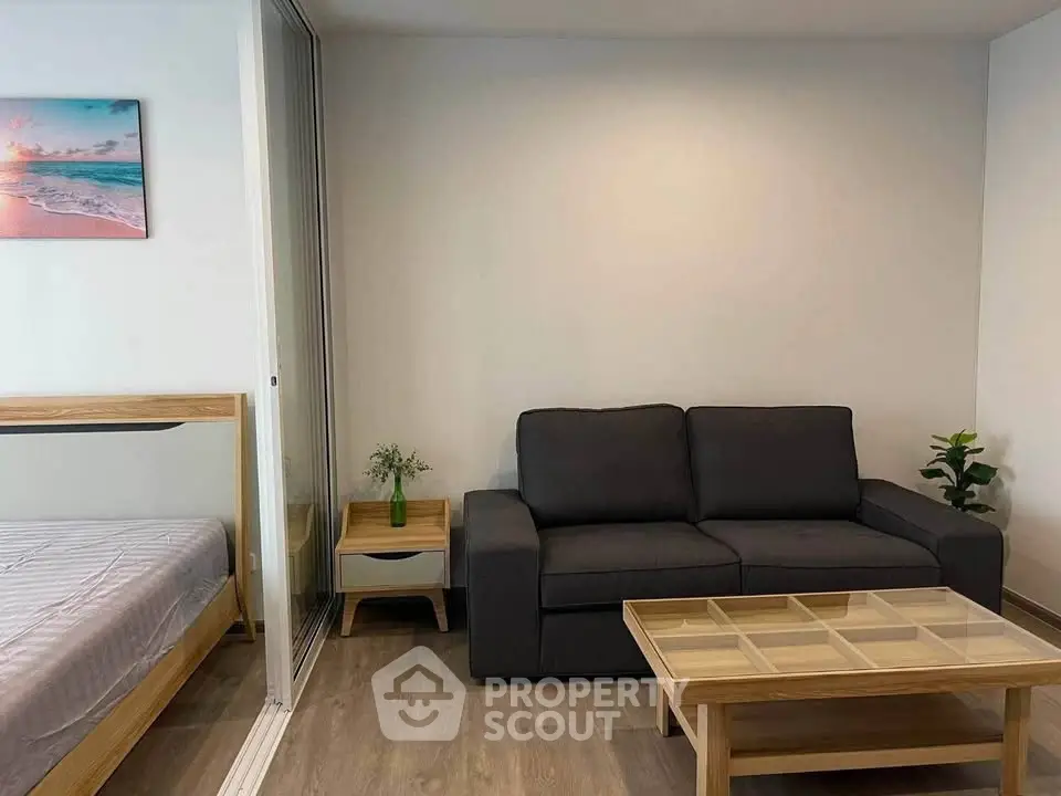 Modern living room with sleek sofa and glass coffee table, adjacent to a cozy bedroom separated by a glass partition.