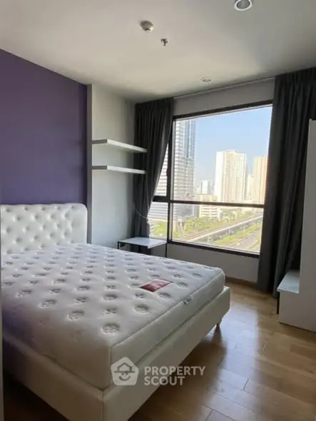 Modern bedroom with large window offering city view, featuring a plush bed and wooden flooring.