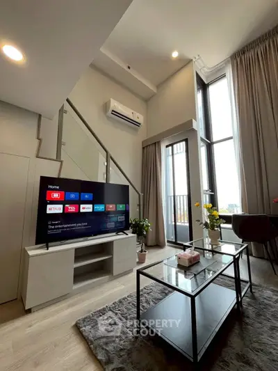 Modern living room with high ceiling and large windows, featuring a sleek TV setup and stylish decor.