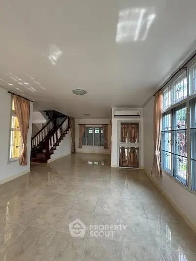 Spacious living room with large windows and elegant curtains, featuring a staircase and tiled flooring.
