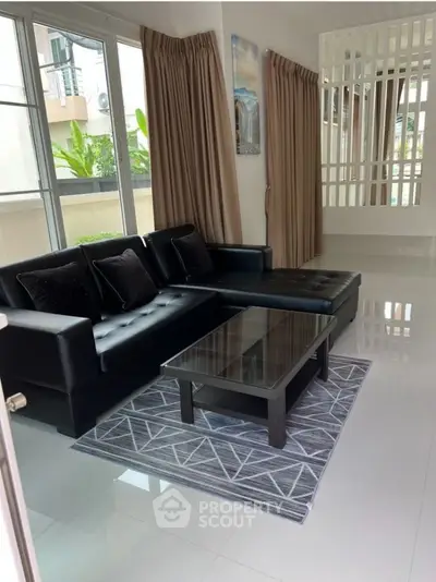 Modern living room with sleek black sofa and glass coffee table, featuring large windows and elegant curtains.