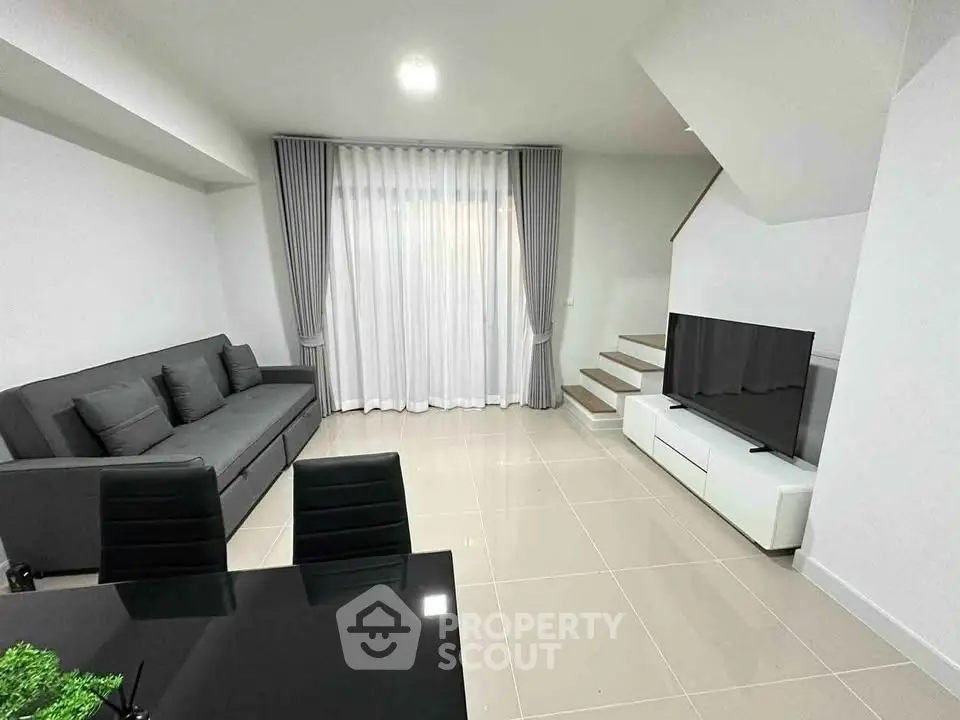 Modern living room with sleek furniture and staircase, featuring a large TV and elegant curtains.