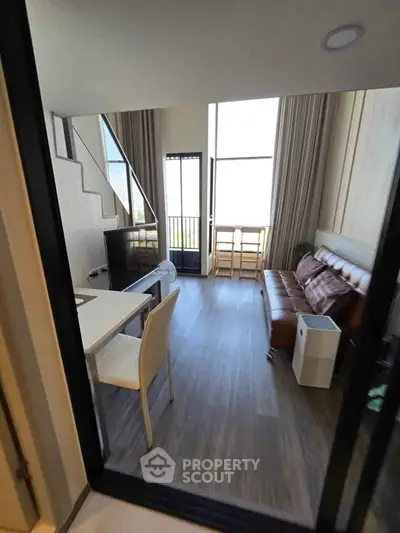 Modern loft-style living room with high ceilings and large windows, featuring a cozy leather sofa and dining area.