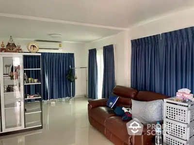 Spacious living room with glossy tiled flooring, large windows draped with blue curtains, and a comfortable brown leather sofa set.