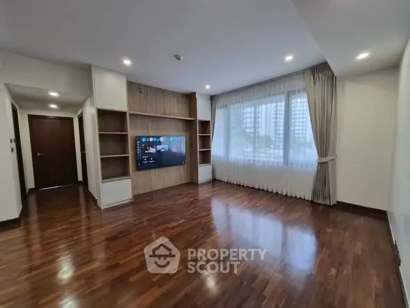 Spacious living room with wooden flooring and built-in TV unit, large window with city view.