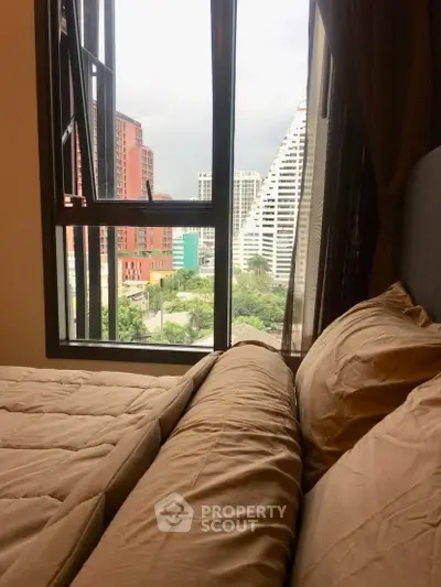 Cozy bedroom with city view through large window, featuring modern bedding and urban skyline.