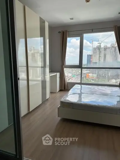 Modern bedroom with large window offering city view, featuring sleek wardrobe and wooden flooring.
