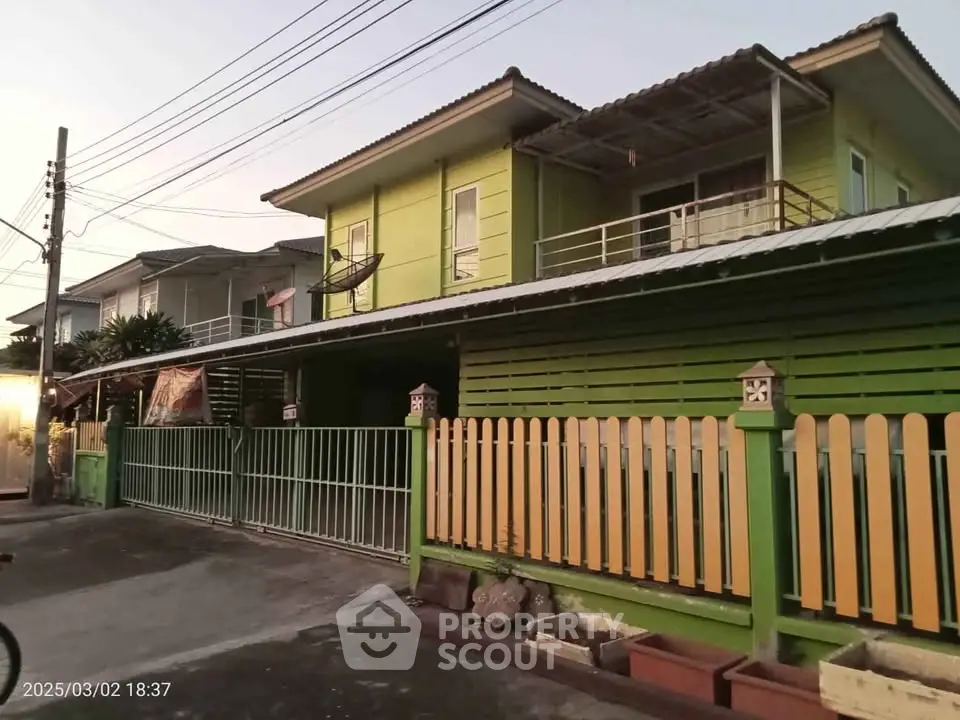 Charming two-story green house with balcony and gated entrance in a serene neighborhood.