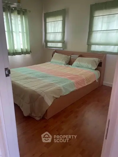 Cozy bedroom with wooden flooring and pastel bedding, featuring natural light through green curtains.