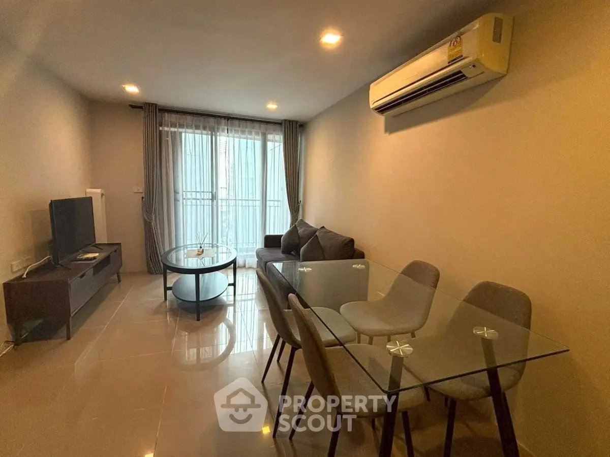 Modern living room with glass dining table and cozy seating area, featuring natural light from large windows.