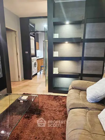 Modern living room with cozy sofa, glass coffee table, and built-in shelving, featuring a glimpse of a compact kitchen.