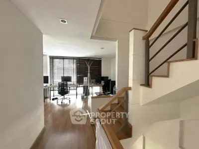Modern home office with wooden flooring and staircase, featuring multiple workstations and natural light.