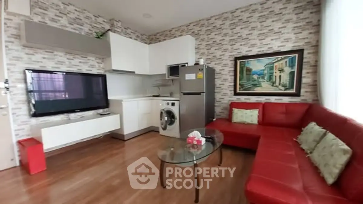 Modern living room with red sofa, wall-mounted TV, and compact kitchen featuring washing machine and fridge.