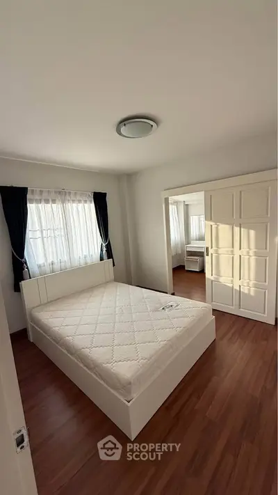 Spacious bedroom with wooden flooring and natural light, featuring a comfortable bed and elegant curtains.
