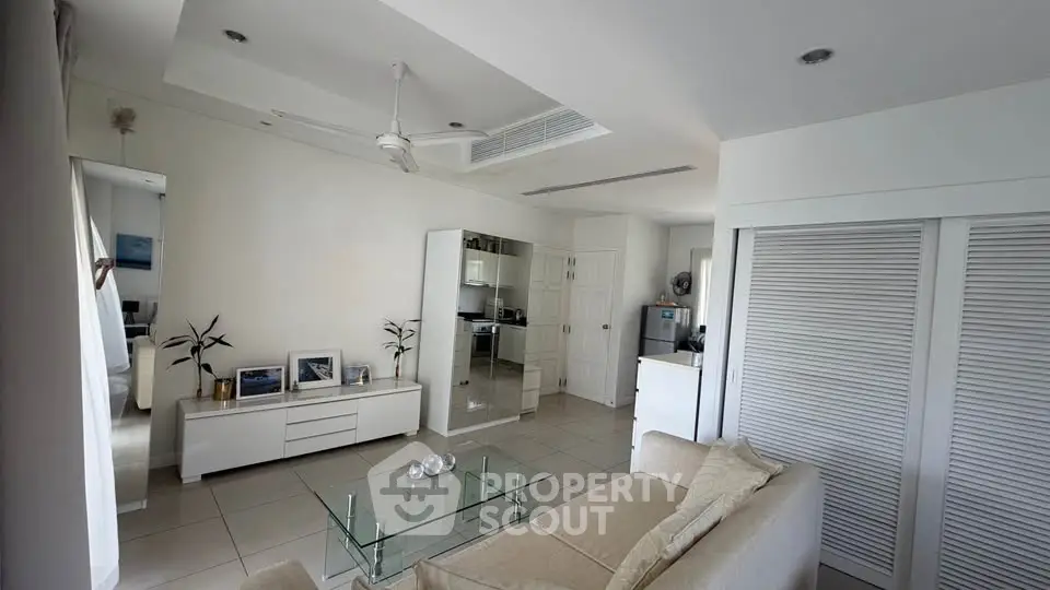 Modern living room with sleek white furniture and glass coffee table, open layout to kitchen area.