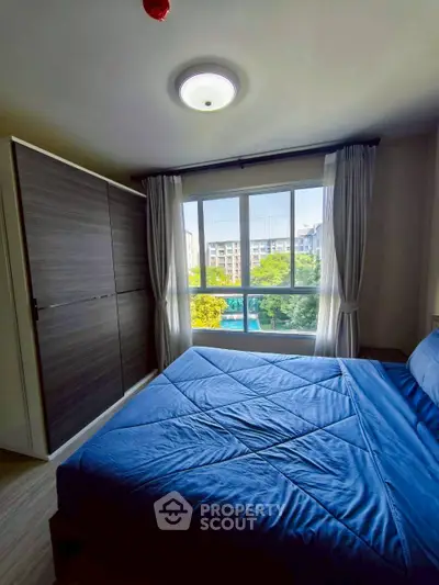 Cozy bedroom with large window and blue bedding, offering a serene view of greenery.