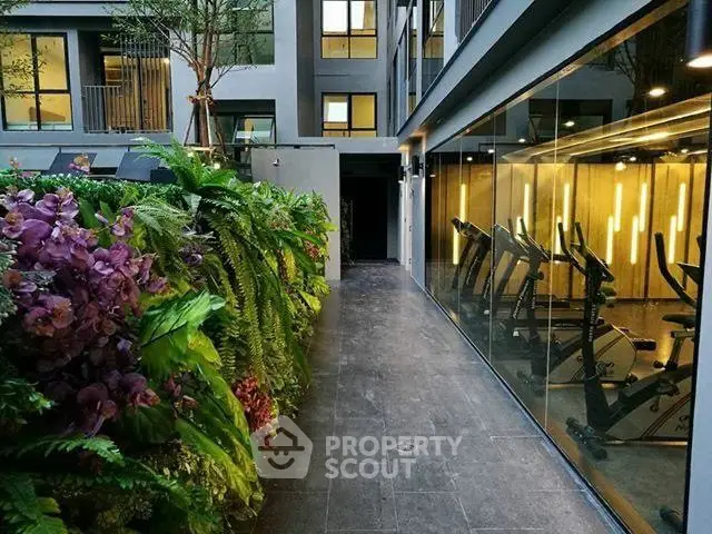 Modern apartment building with lush garden and glass-walled gym showcasing exercise bikes.