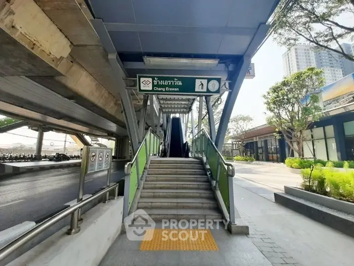 Modern urban entrance with escalator and signage, surrounded by greenery and cityscape.