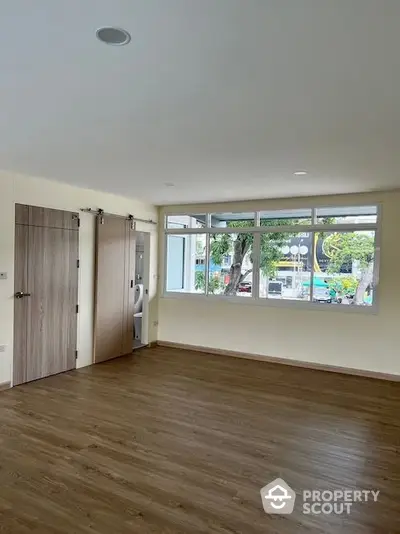 Spacious and well-lit living space with large windows offering an urban view, modern wood flooring, and sliding doors leading to a cozy bedroom.