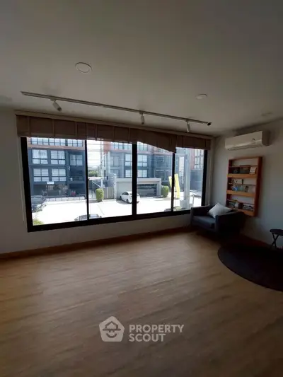 Spacious living room with large windows offering a view of modern buildings, featuring wooden flooring and cozy seating.
