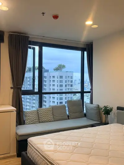 Modern bedroom with large window offering city view, cozy seating, and stylish decor.