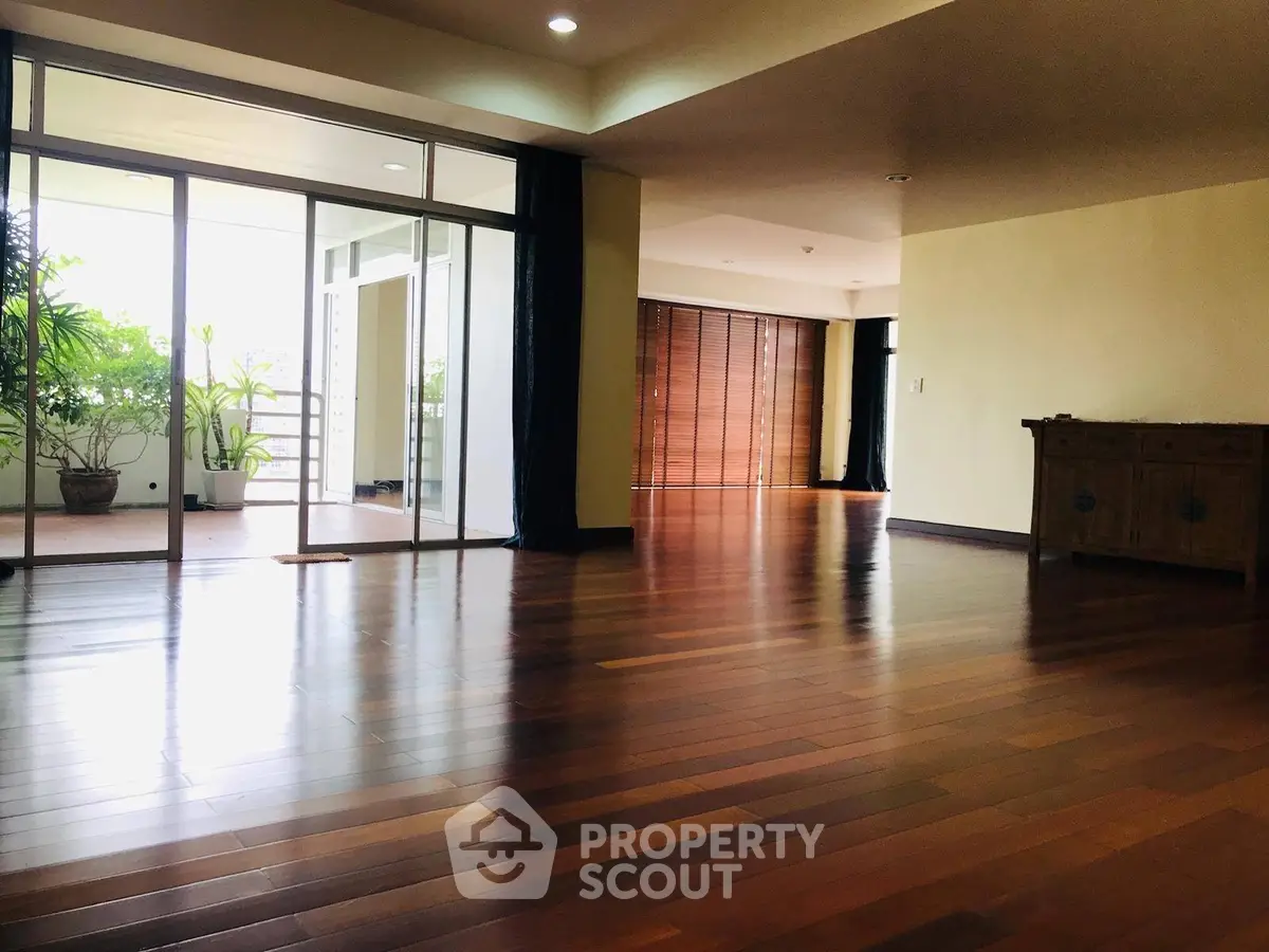 Spacious living room with wooden flooring and large windows opening to a balcony with greenery.