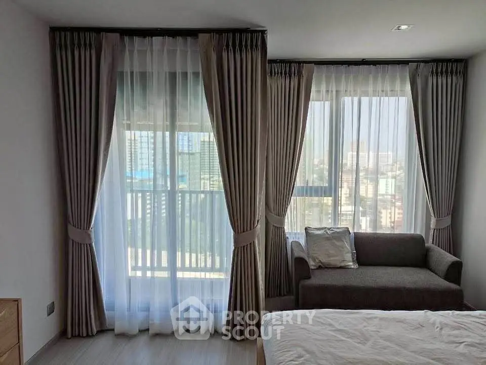 Spacious bedroom with large windows and city view, featuring elegant curtains and cozy seating area.