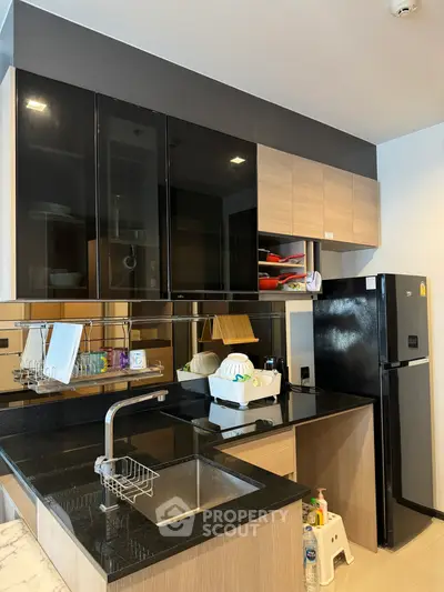 Modern kitchen with sleek black countertops and wooden cabinets, featuring a double-door fridge and stainless steel sink.