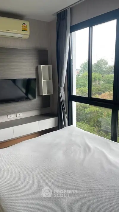 Modern bedroom with large window and wall-mounted TV, offering a serene view of greenery.