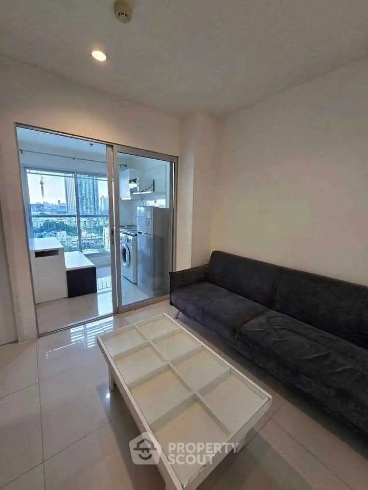 Modern living room with sleek sofa and glass coffee table, adjacent to a bright kitchen with city view.