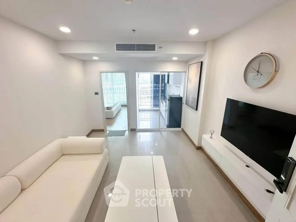 Modern living room with sleek white furniture and wall-mounted TV, leading to a bright balcony view.
