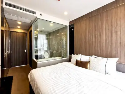 Modern bedroom with glass wall view into luxurious bathroom, featuring elegant wood paneling and plush bedding.