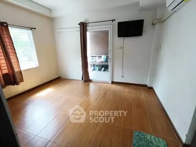 Spacious room with wooden flooring, wall-mounted TV, and large window for natural light.