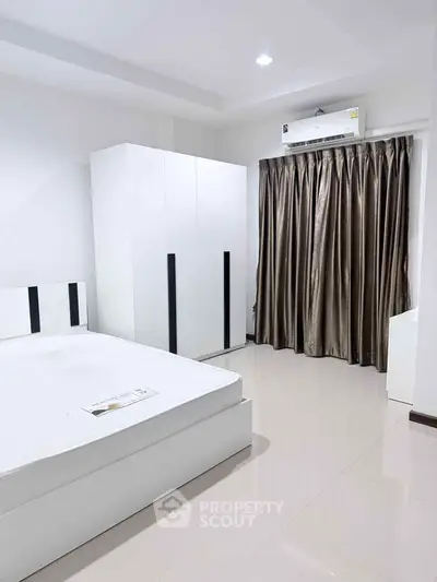 Modern bedroom with sleek white furniture and air conditioning, featuring a minimalist design and ample natural light.
