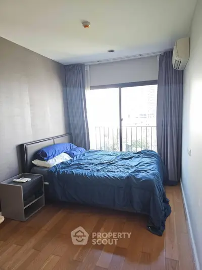 Cozy bedroom with wooden flooring and large window offering natural light and city view.