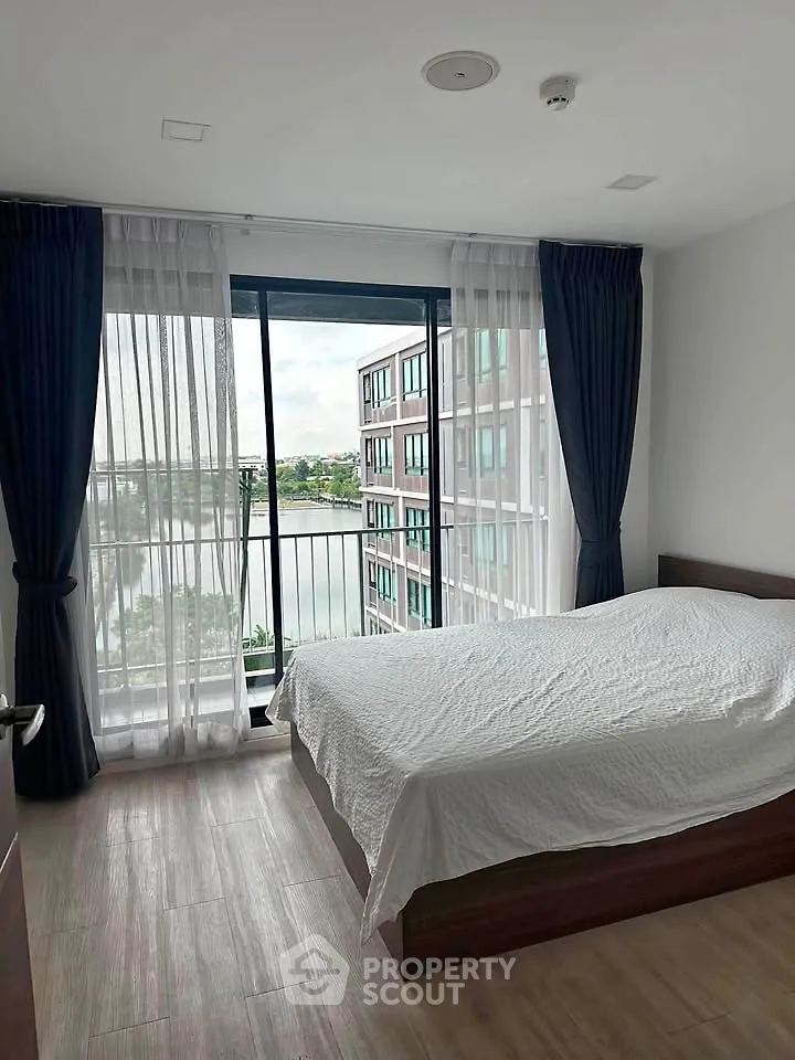 Spacious bedroom with large window offering stunning view of modern building and serene water.