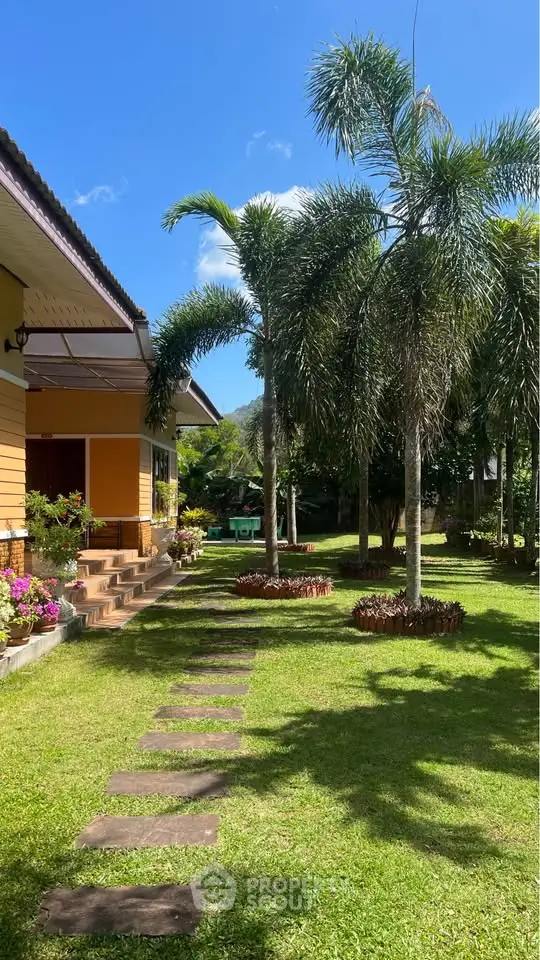 Charming tropical garden with palm trees and a vibrant yellow house under a clear blue sky.
