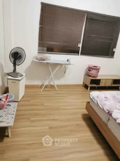 Cozy bedroom with wooden flooring, bed, fan, and ironing board, perfect for comfortable living.