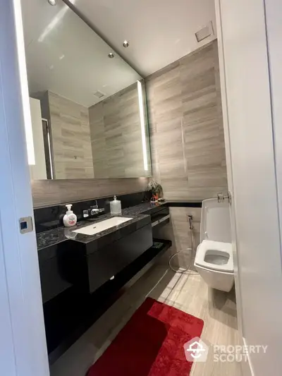 Modern bathroom with sleek design, featuring a large mirror, elegant vanity with dark countertop, and stylish tiling throughout, complemented by a red accent rug.