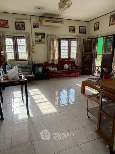 Spacious living room with natural light and tiled flooring, featuring comfortable seating and air conditioning.