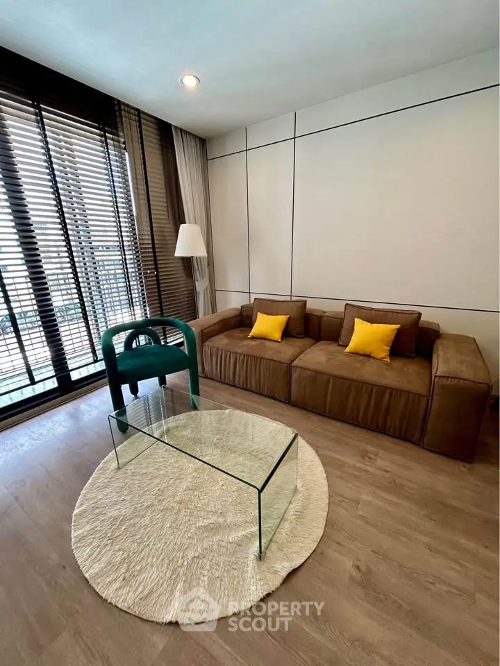 Modern living room with stylish brown sofa, glass coffee table, and floor-to-ceiling windows with blinds.