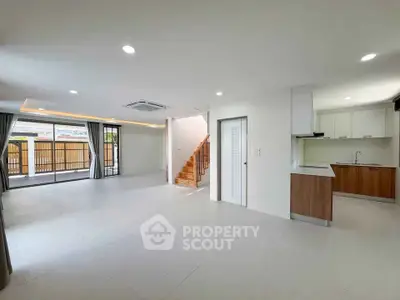 Spacious open-plan living room with modern kitchen and staircase, featuring large windows and ample natural light.