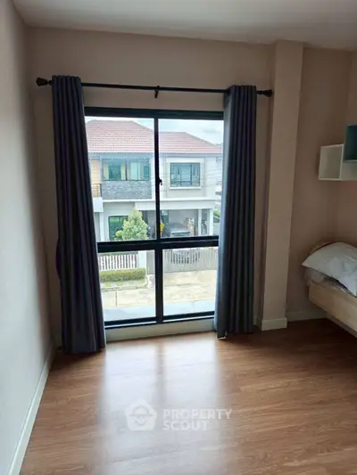 Modern bedroom with large window and wooden flooring, offering a view of suburban homes.