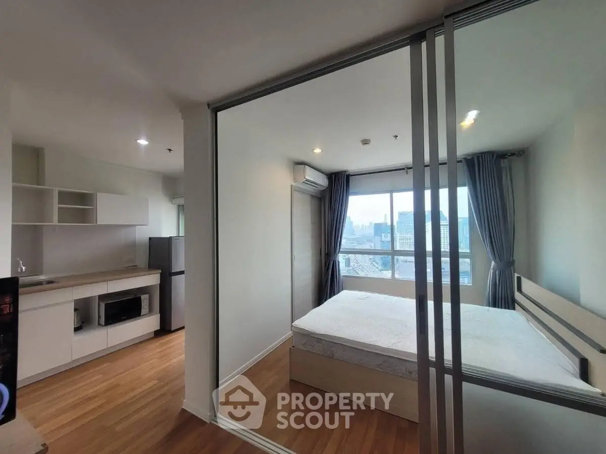 Modern apartment with glass-walled bedroom and city view, featuring sleek kitchen and wooden flooring.