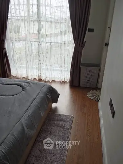 Cozy bedroom with wooden flooring and large window with sheer curtains.